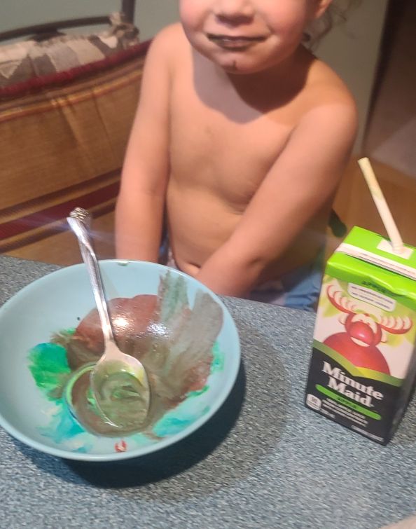 A toddler sitting at a kitchen island in front of an apple juice box and an empty dish of food-coloured vanilla ice cream