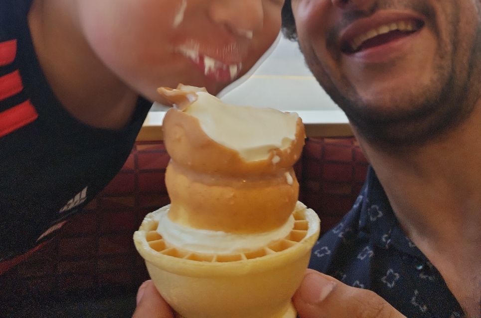 A toddler stuffing his face with dad's dipped soft serve ice cream cone.