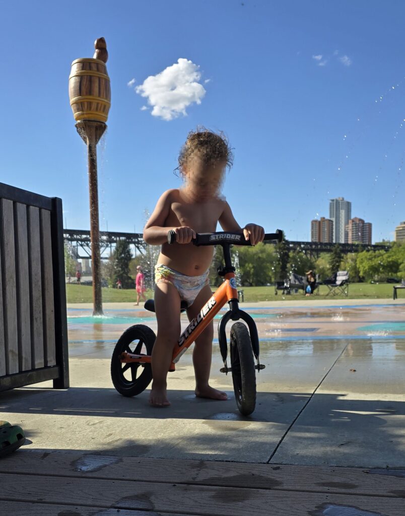A toddler on a Strider balance bike at a spray park, moments before getting a helmet slapped on.