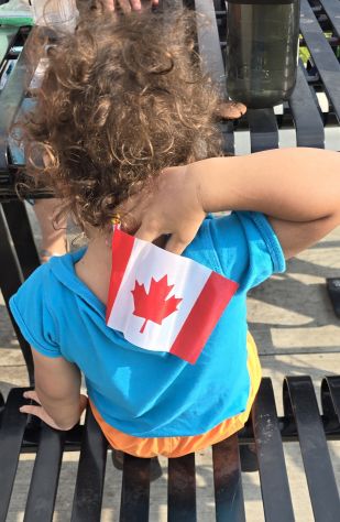 A rear view of a seated toddler trying to move a Canad flag placed in the back of their shirt.
