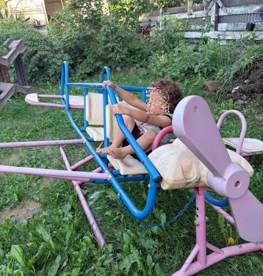 A toddler in a lush back yard on a stray piece of playground equipment shaped like an airplane.